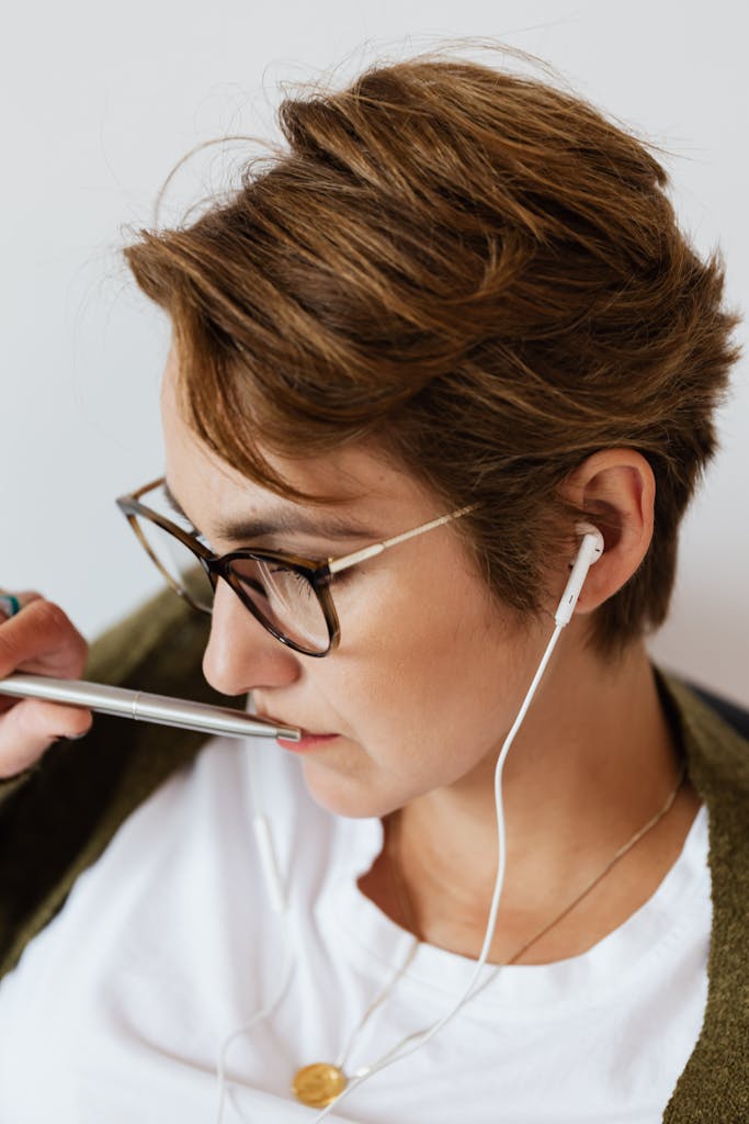 Focused female in casual clothes with stylish necklace and eyeglasses listening to podcast in earphones and touching lips with silver pen against white wall